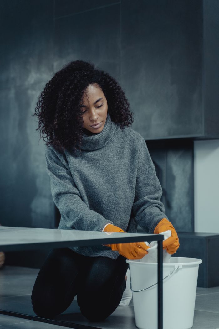 hero-img African American woman in gray sweater cleaning with a pail and gloves at home.